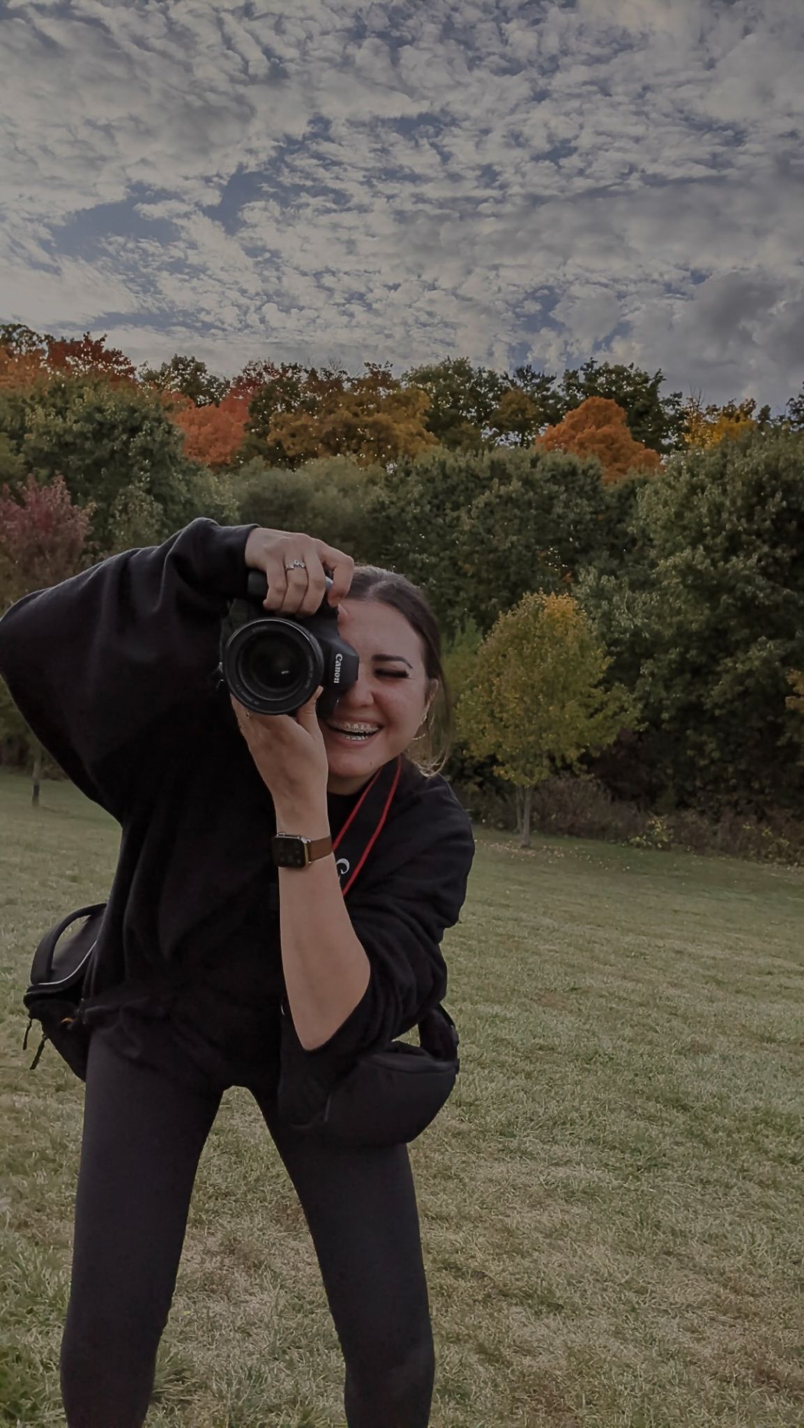 Elopement Photographer located in Ontario in black clothing takes photos in an autumn field with cloudy skies overhead.