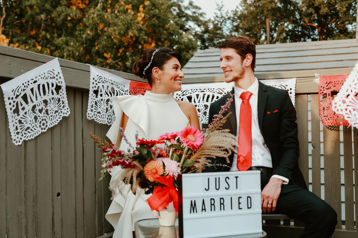 Newlyweds sit together on bench with Just Married sign and papel picado banners at outdoor wedding celebration.