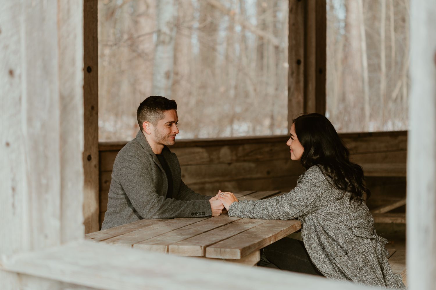 Two people sit in conversation at a rustic wooden table in an abandoned cabin.