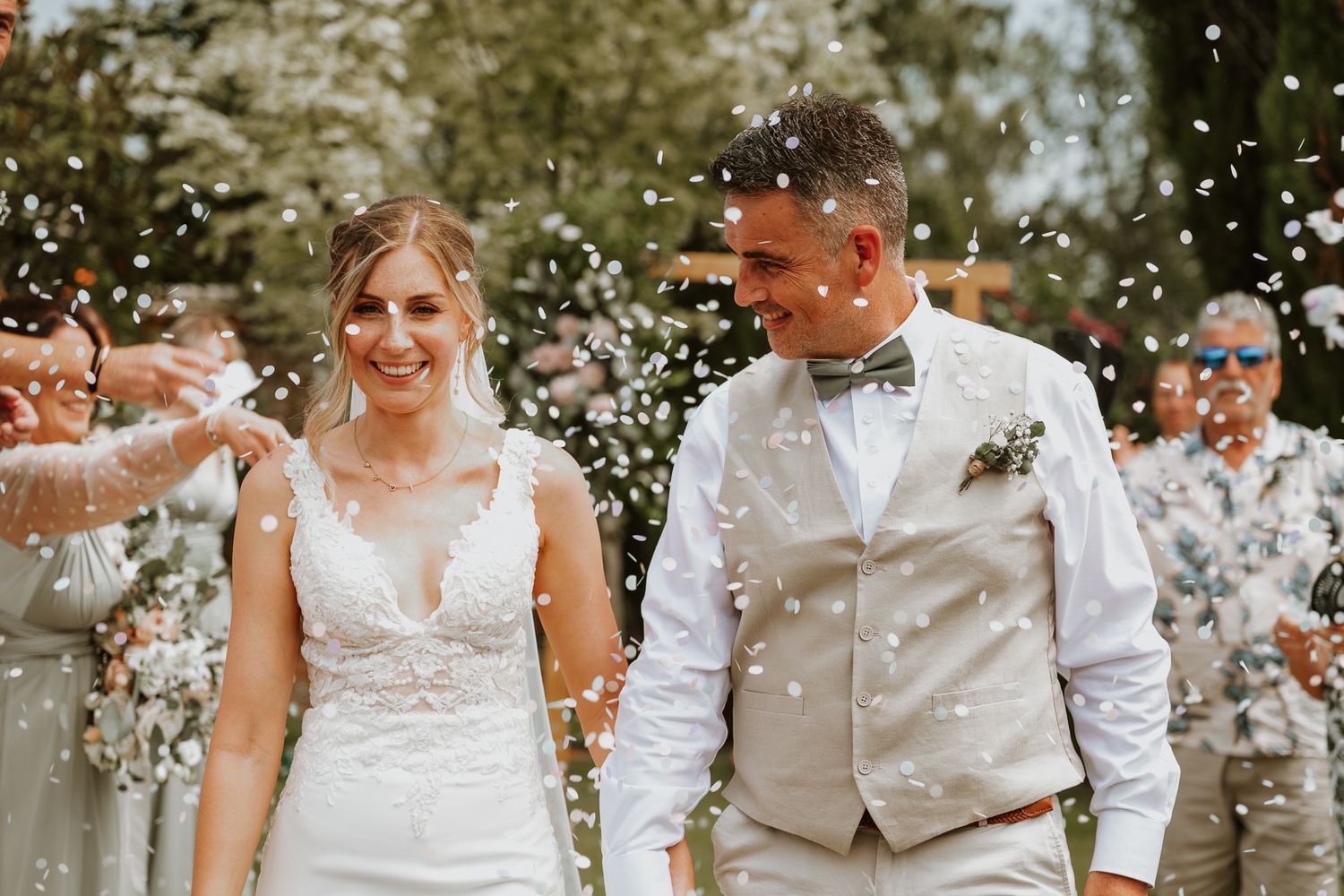A joyful wedding couple walks through white flower petals being showered by guests during their outdoor celebration.