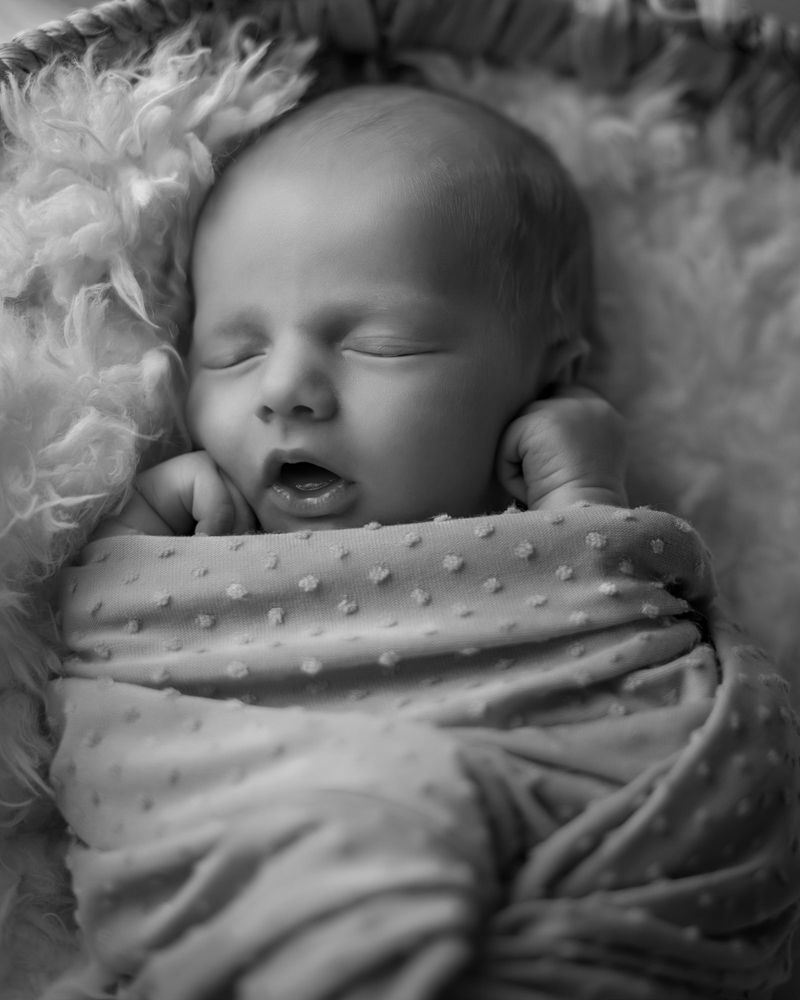 Black and white newborn baby sleeping peacefully, swaddled in dotted blanket on fluffy white fur background.