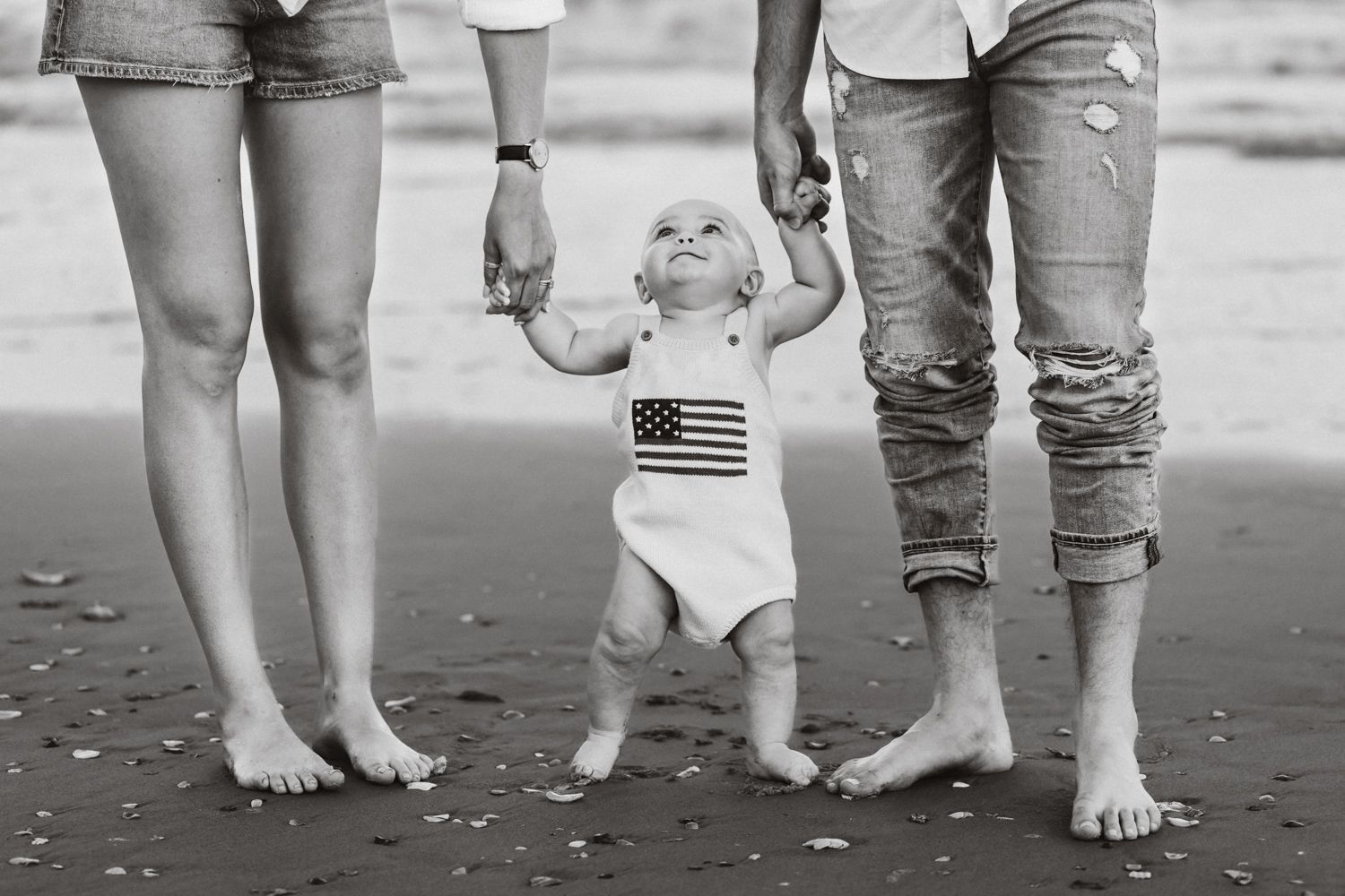 Black and white photo of a baby in American flag onesie learning to walk on beach held by two adults.