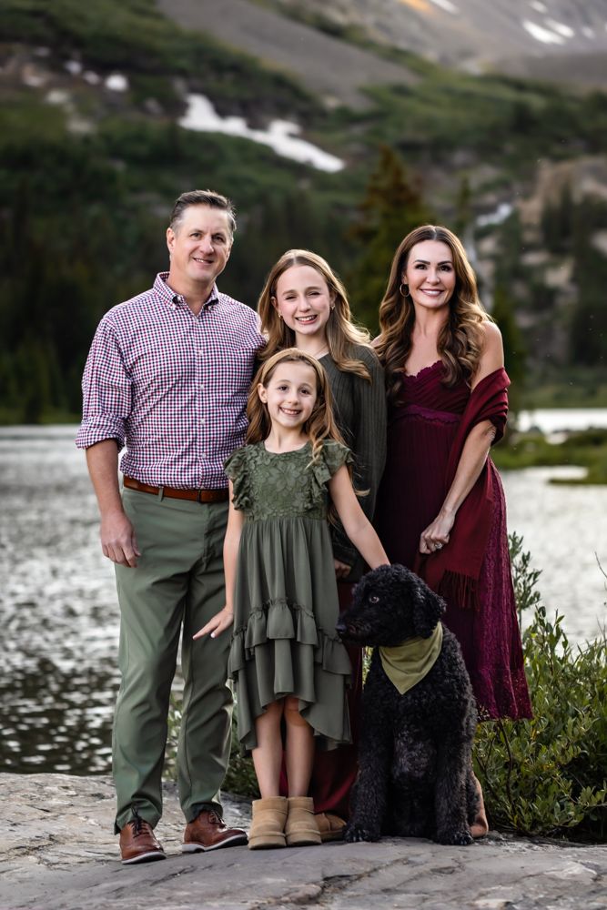 Family of four posing with black dog near mountain lake, wearing coordinated green and burgundy outfits.