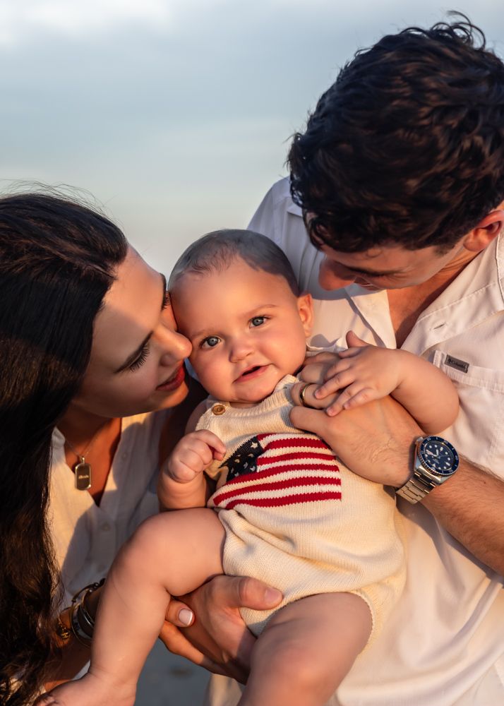 Parents kissing their smiling baby wearing an American flag onesie outdoors at golden hour.