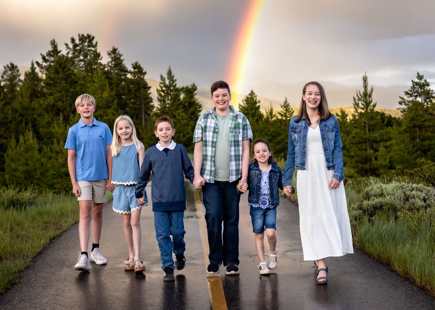 Six children walk hand-in-hand on a road with a vibrant rainbow and lush green trees in the background.