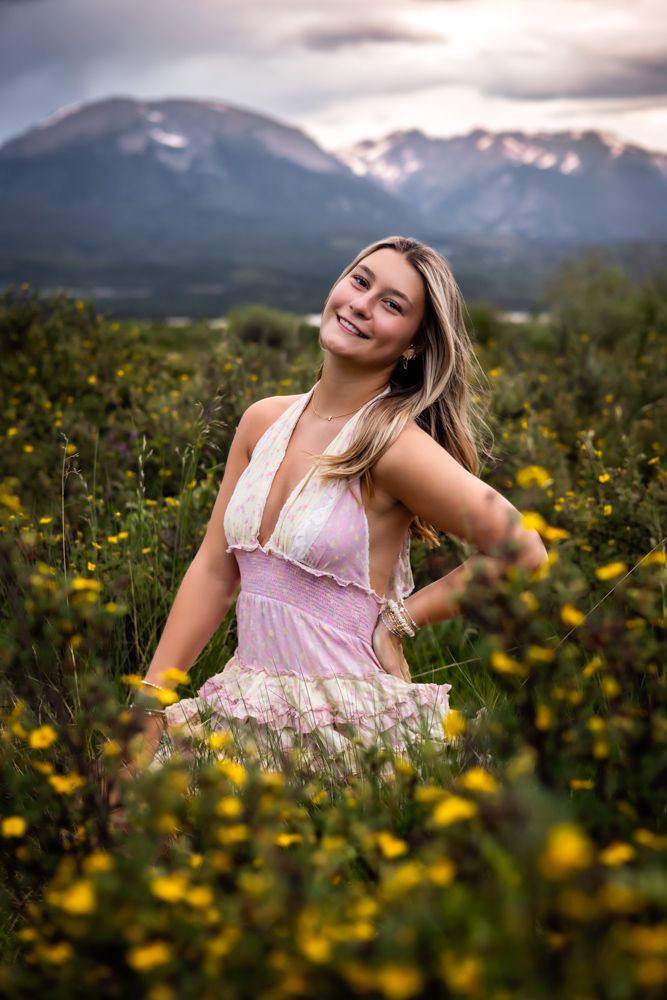 Young blonde woman in pink floral dress smiling in wildflower field with snow-capped mountains at sunset.