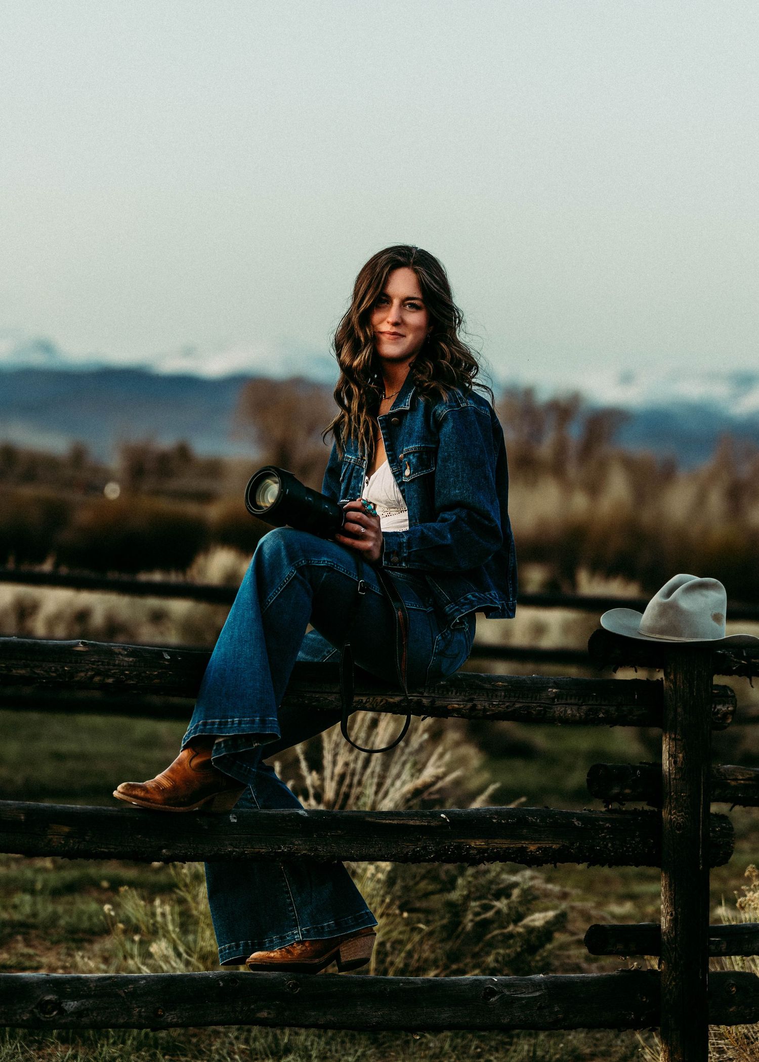 Cowgirl in denim sits casually on wooden fence at dusk with mountains in background.