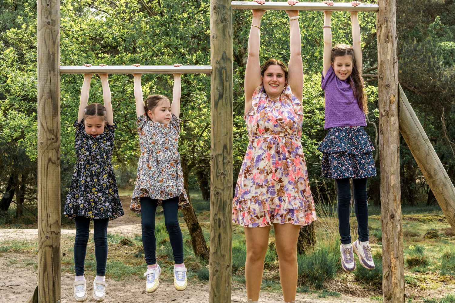 Four children hanging from wooden playground equipment in a forest setting.