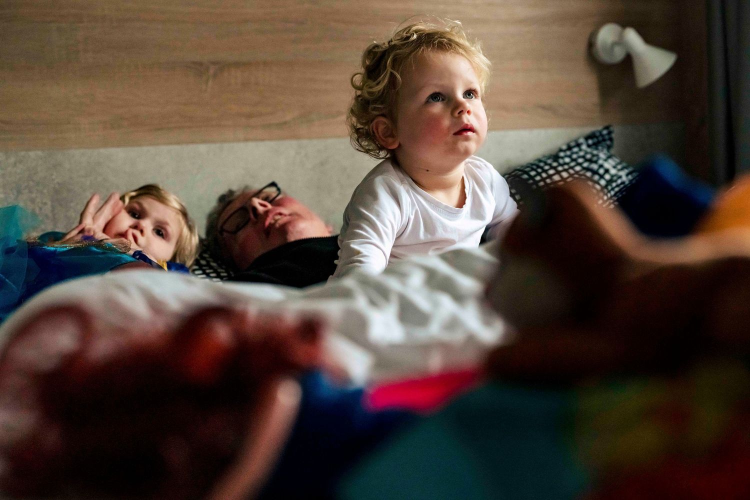 Young child gazing upward while others rest on bed in natural lighting.