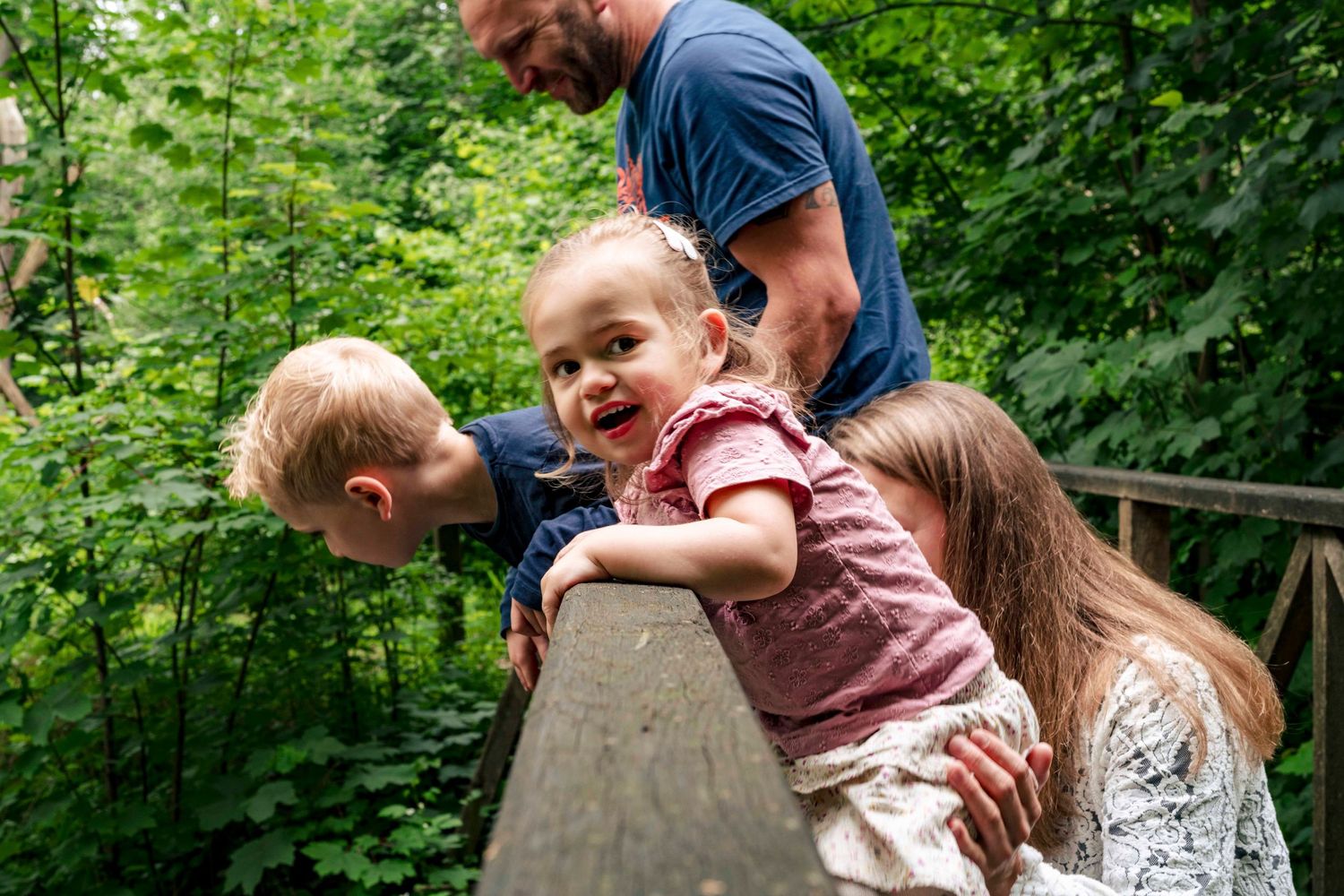 Children laughing while playing on wooden railing in lush green forest setting.