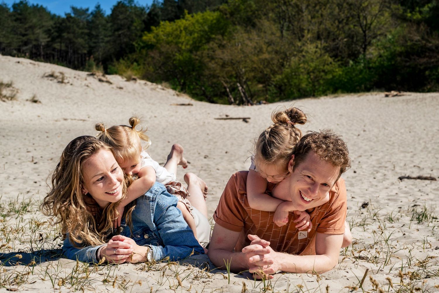 Family lying together on sandy beach with forest backdrop on sunny day.