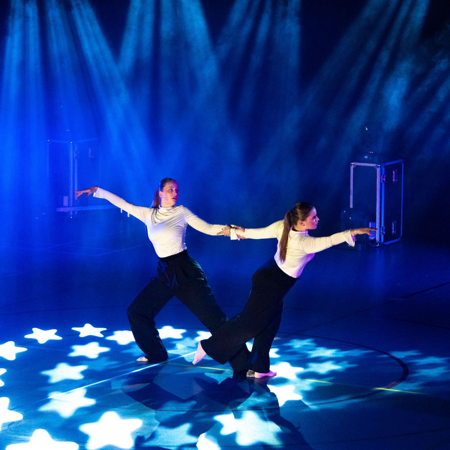 Two dancers in black pants strike a pose on stage with blue lighting and star-shaped floor projections.