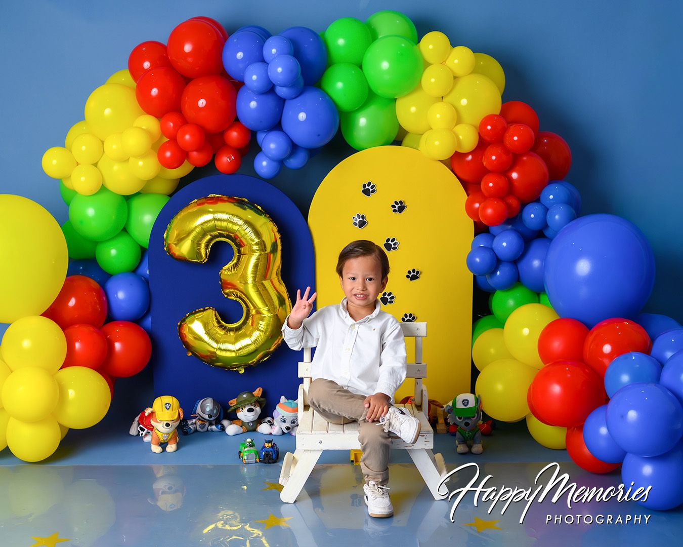 Colorful balloon arch display with red, blue, green and yellow balloons featuring a gold number 3 against blue backdrop.