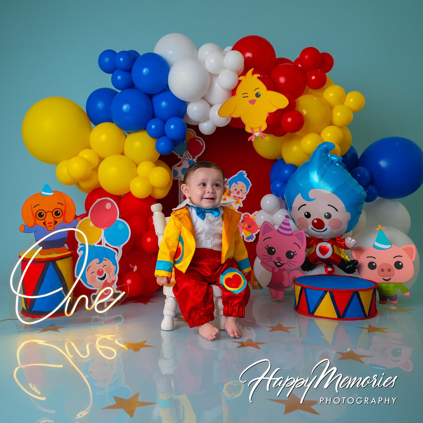 Baby boy celebrates first birthday with colorful circus-themed balloon arch, clown toys, and drum decor by Happy Memories Photography.