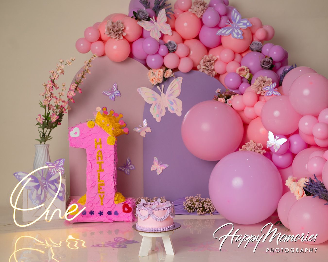 Pink and purple balloon arch with butterflies and floral decorations against a beige wall creates a romantic backdrop.