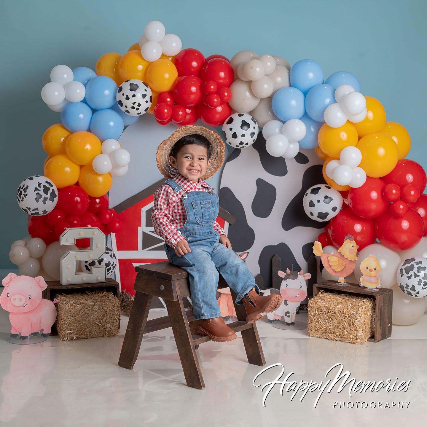 A young boy in overalls and cowboy hat sits on a stool surrounded by colorful farm-themed balloons and hay bales.