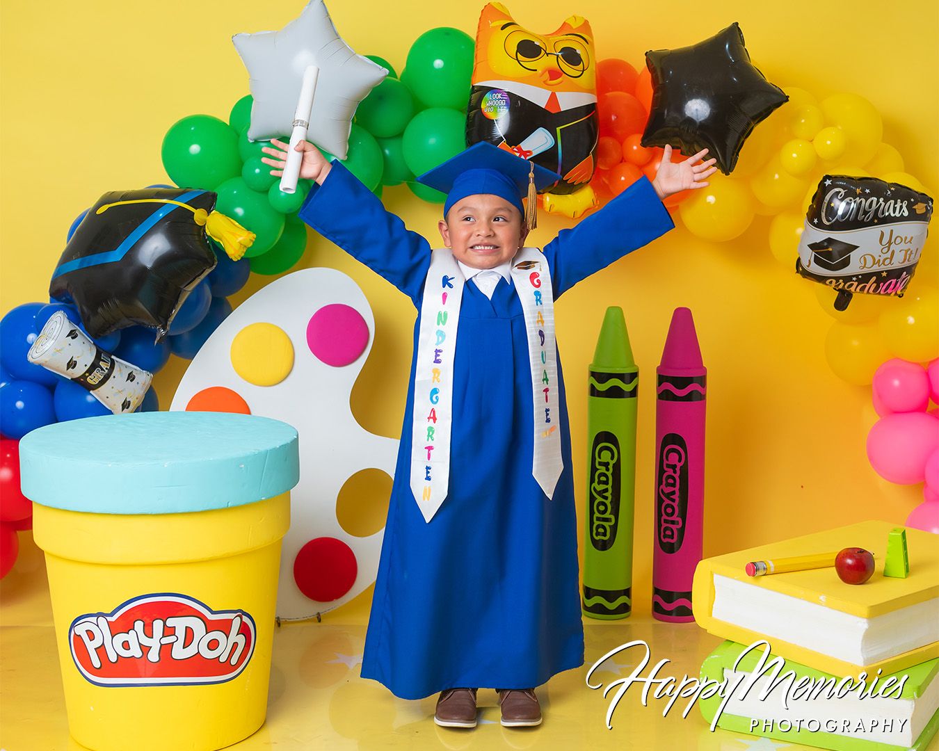 Playful preschool graduation setup with art supplies, blue cap and gown, and rainbow balloon arch.