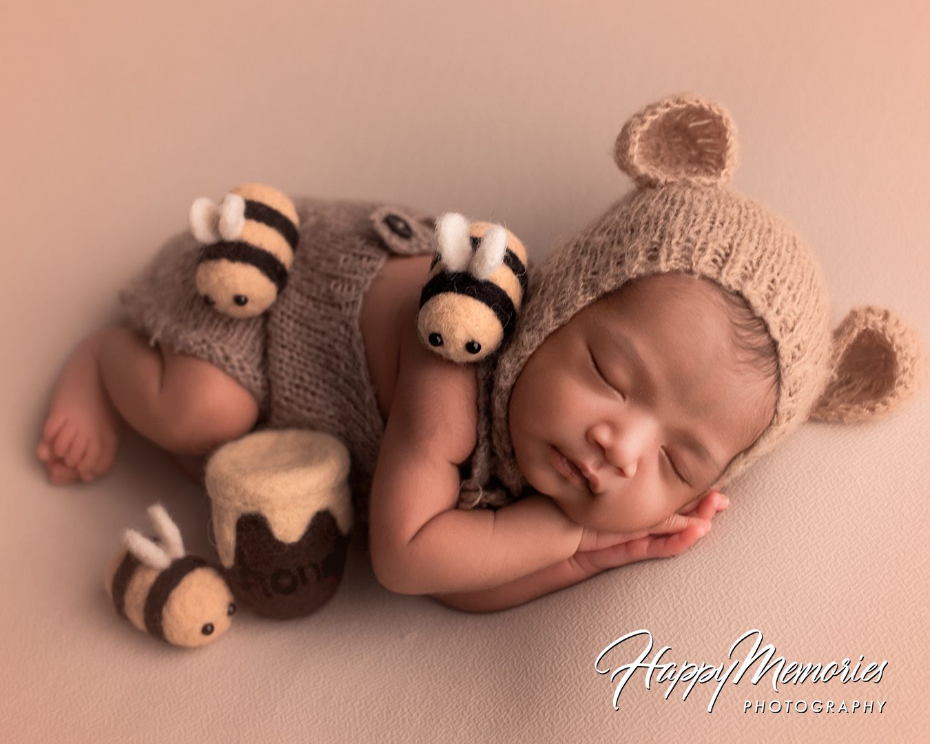 Sleeping newborn wearing knitted bear outfit lying next to plush teddy bear in soft pink studio lighting.