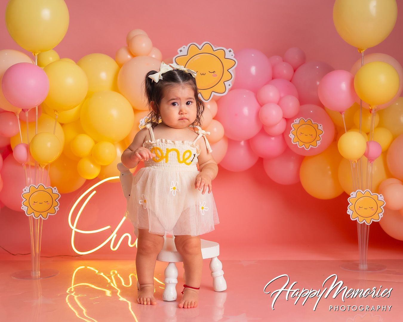 Pink and gold balloon arch decorates a birthday cake smash photo session against a coral backdrop.