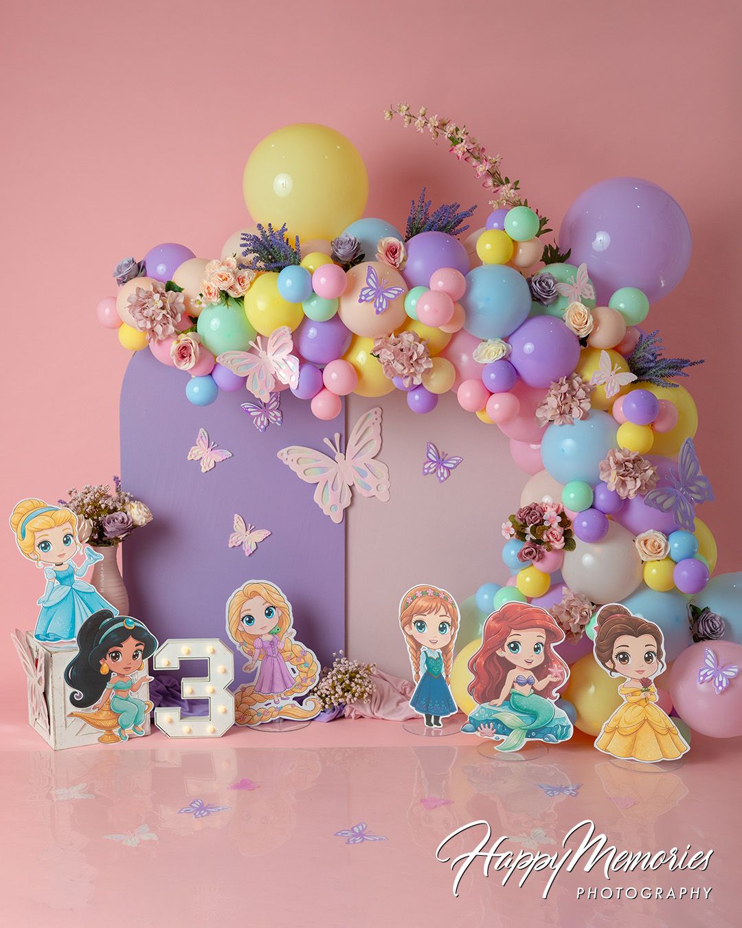 Disney princess birthday cake smash setup with colorful balloon arch, floral decor, and princess cutouts on pink background.