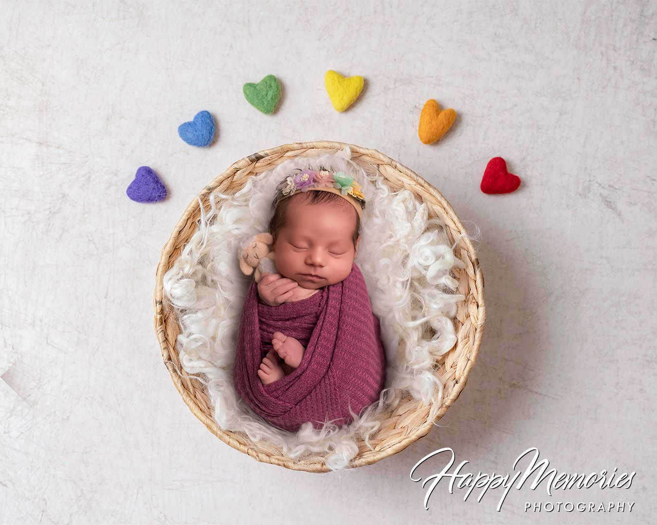 Sleeping newborn in white basket with colorful heart decorations arranged on light background.