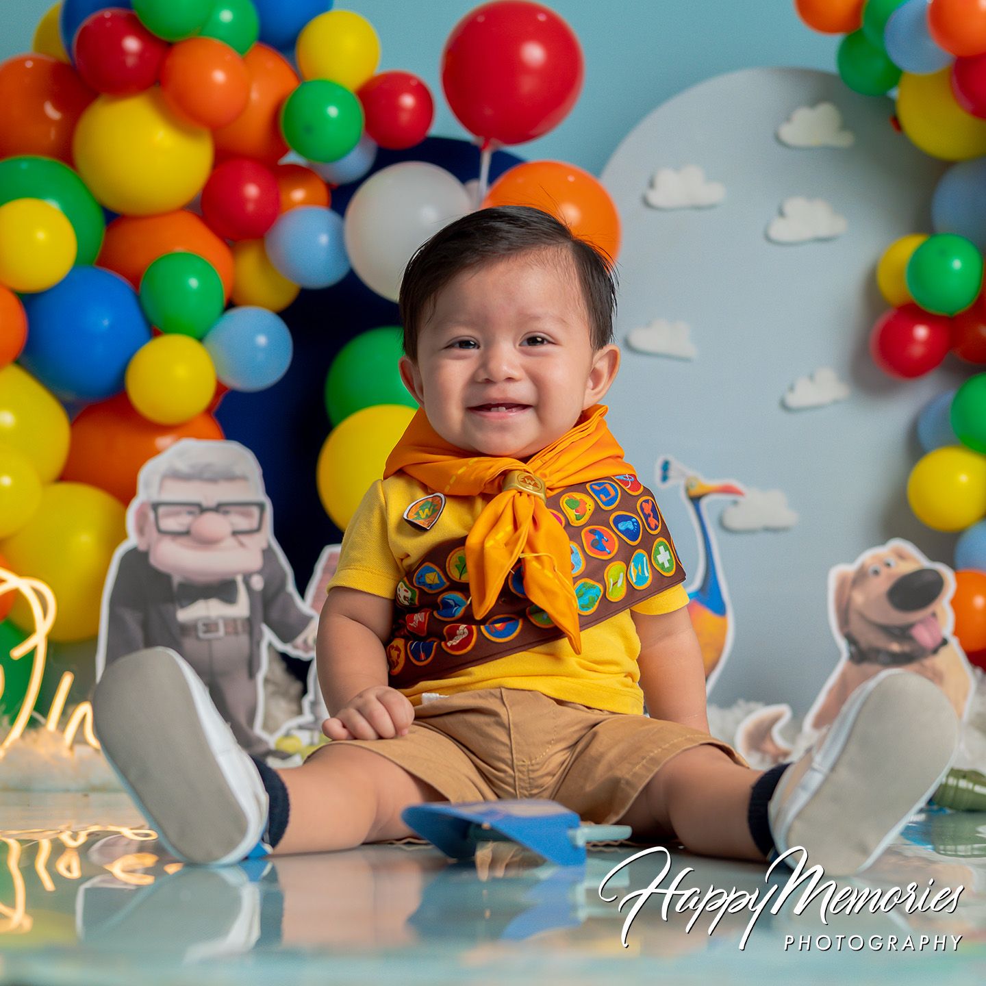 Baby dressed as Russell from Pixar Up, smiling at camera surrounded by colorful balloons and Up-themed backdrop.