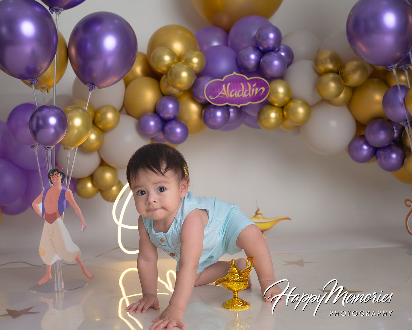 Purple and gold balloon arch decorates a room with a person in light blue sitting near a table.