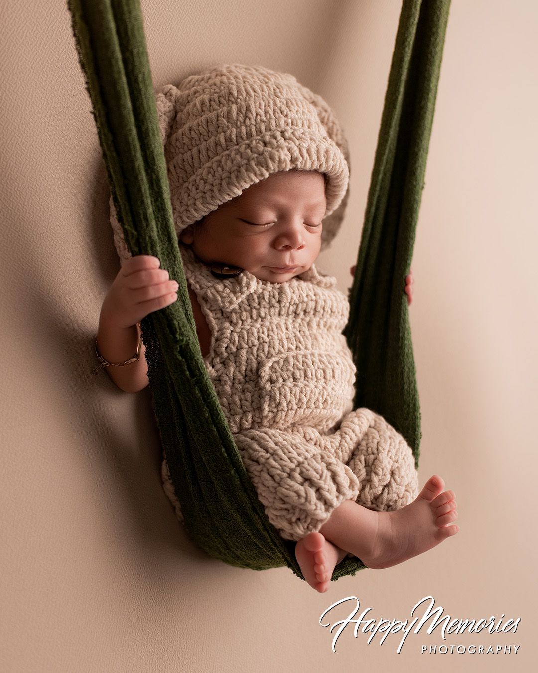 Sleeping newborn in knitted beige outfit rests peacefully in green fabric hammock during professional photo session.