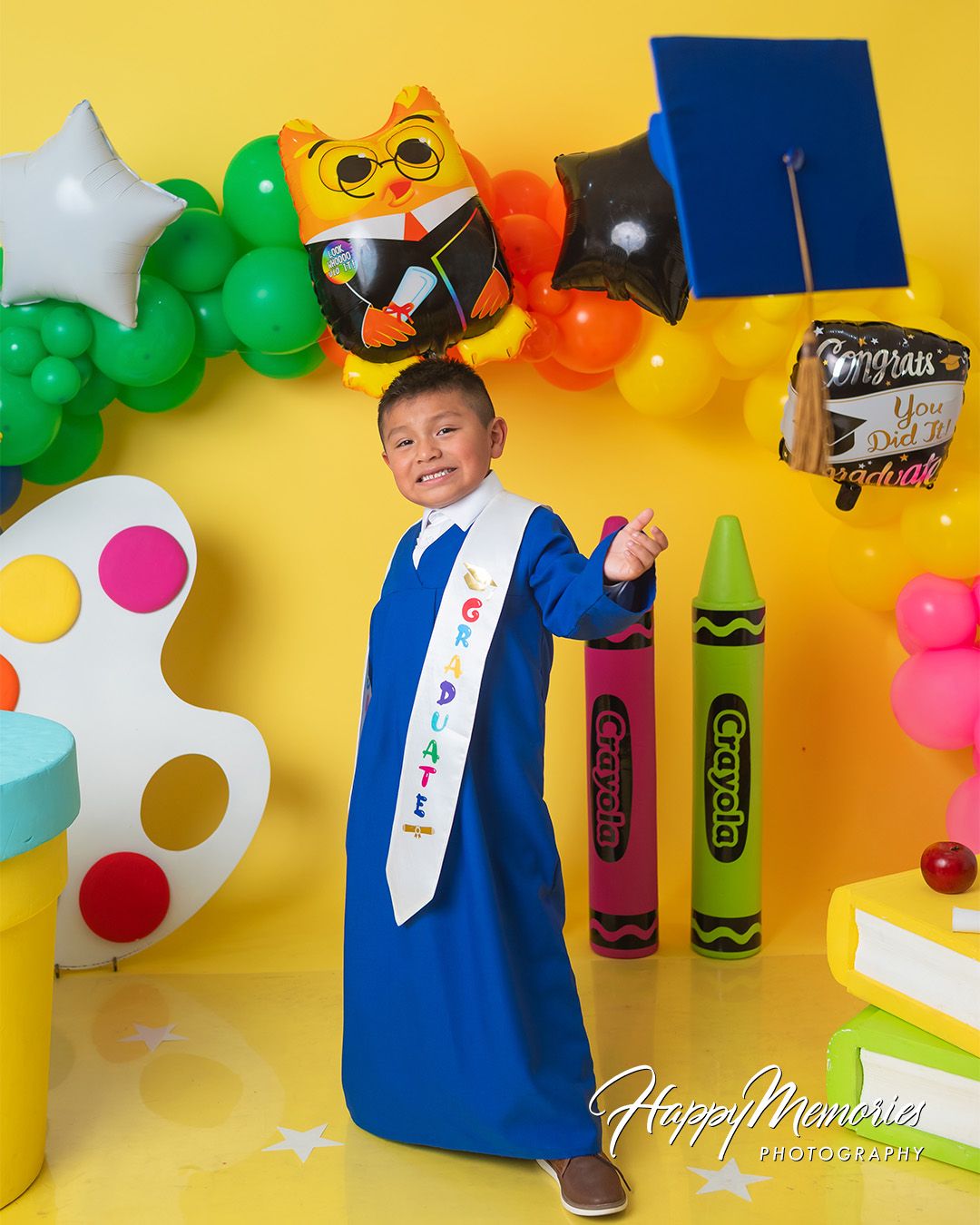 Educational themed graduation photo setup with art supplies and rainbow balloon garland.