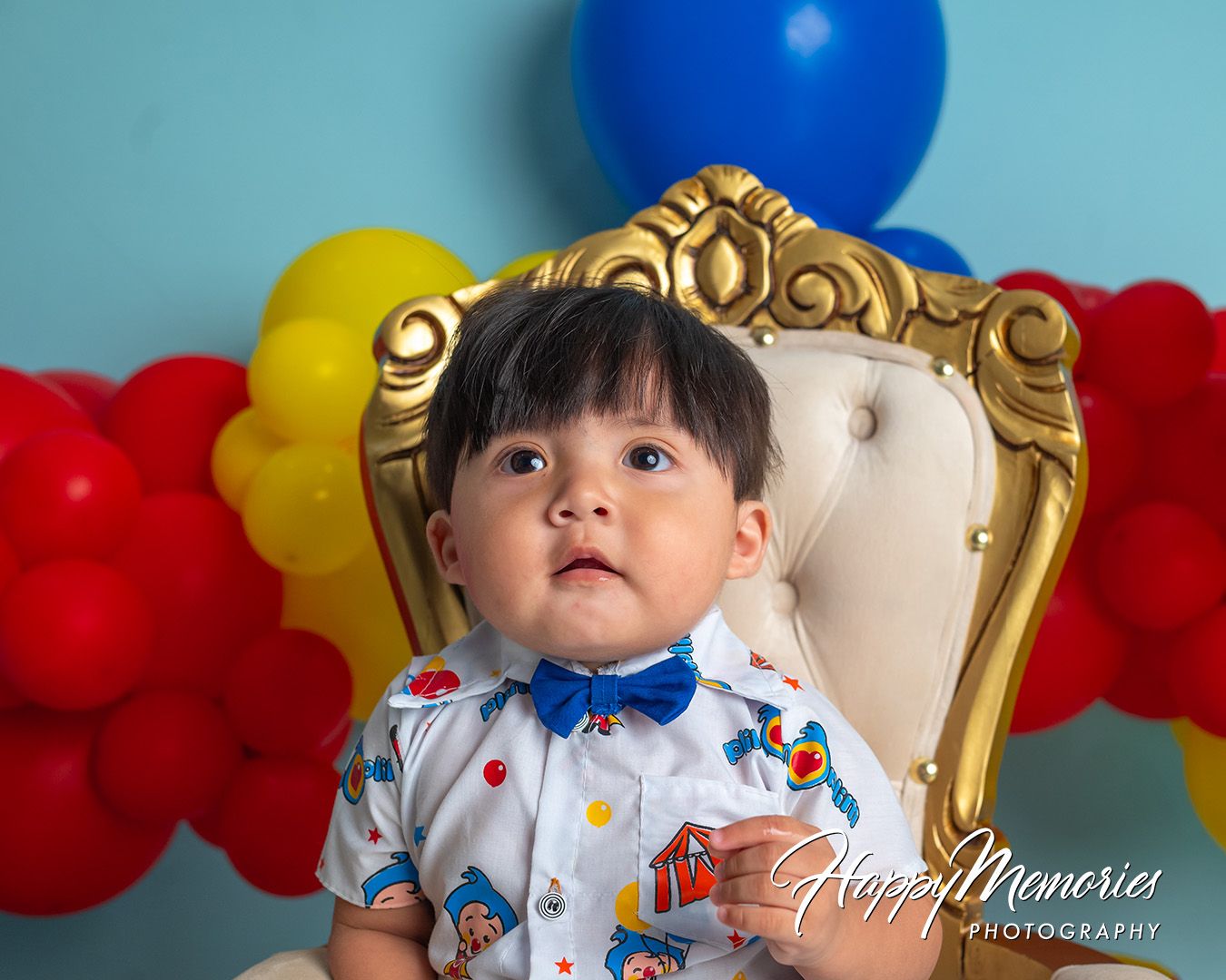 A child sits in an ornate gold throne chair with colorful balloon decorations in red, blue and yellow behind them.