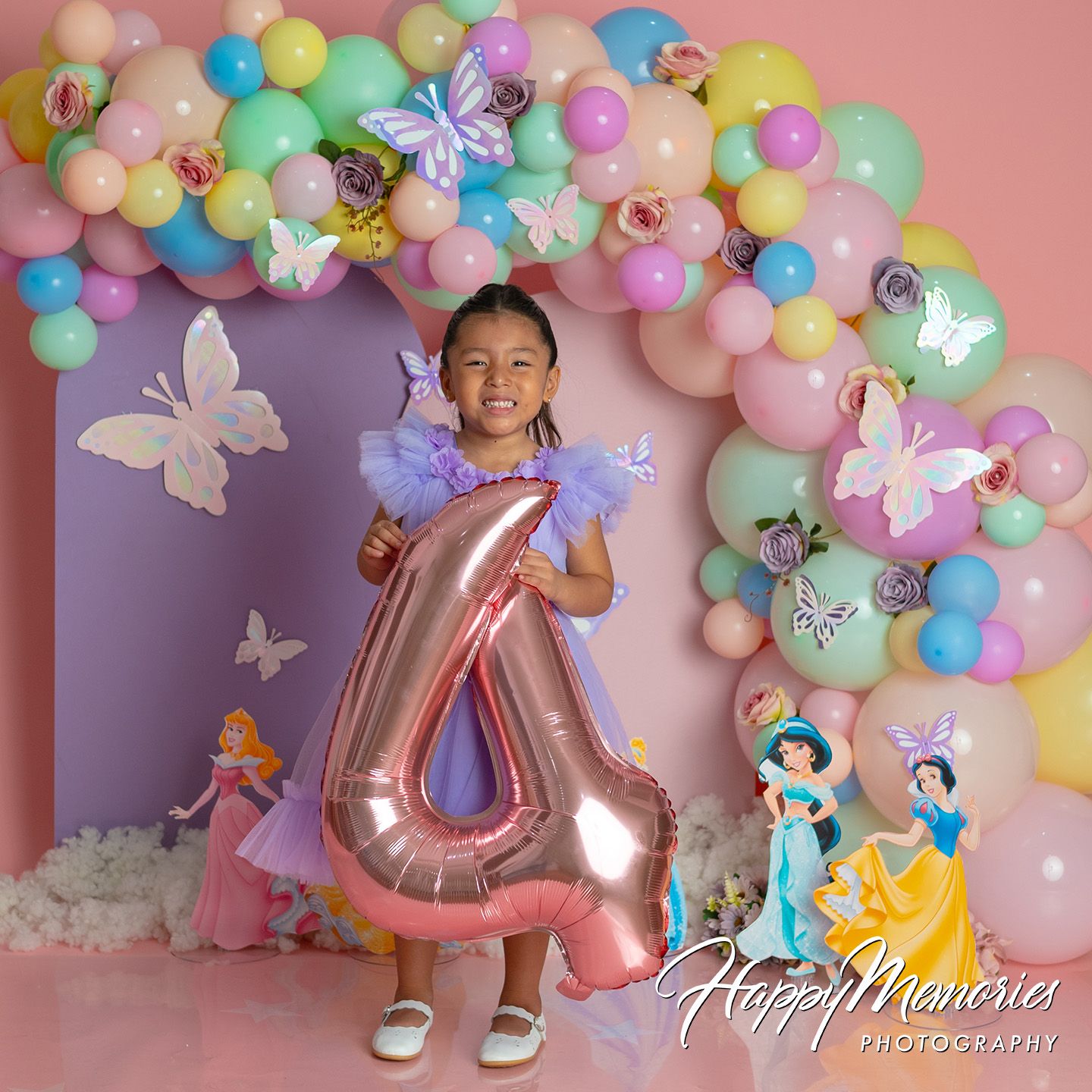 Girl holding number 4 balloon at princess-themed birthday party with colorful balloon arch and butterfly decorations.