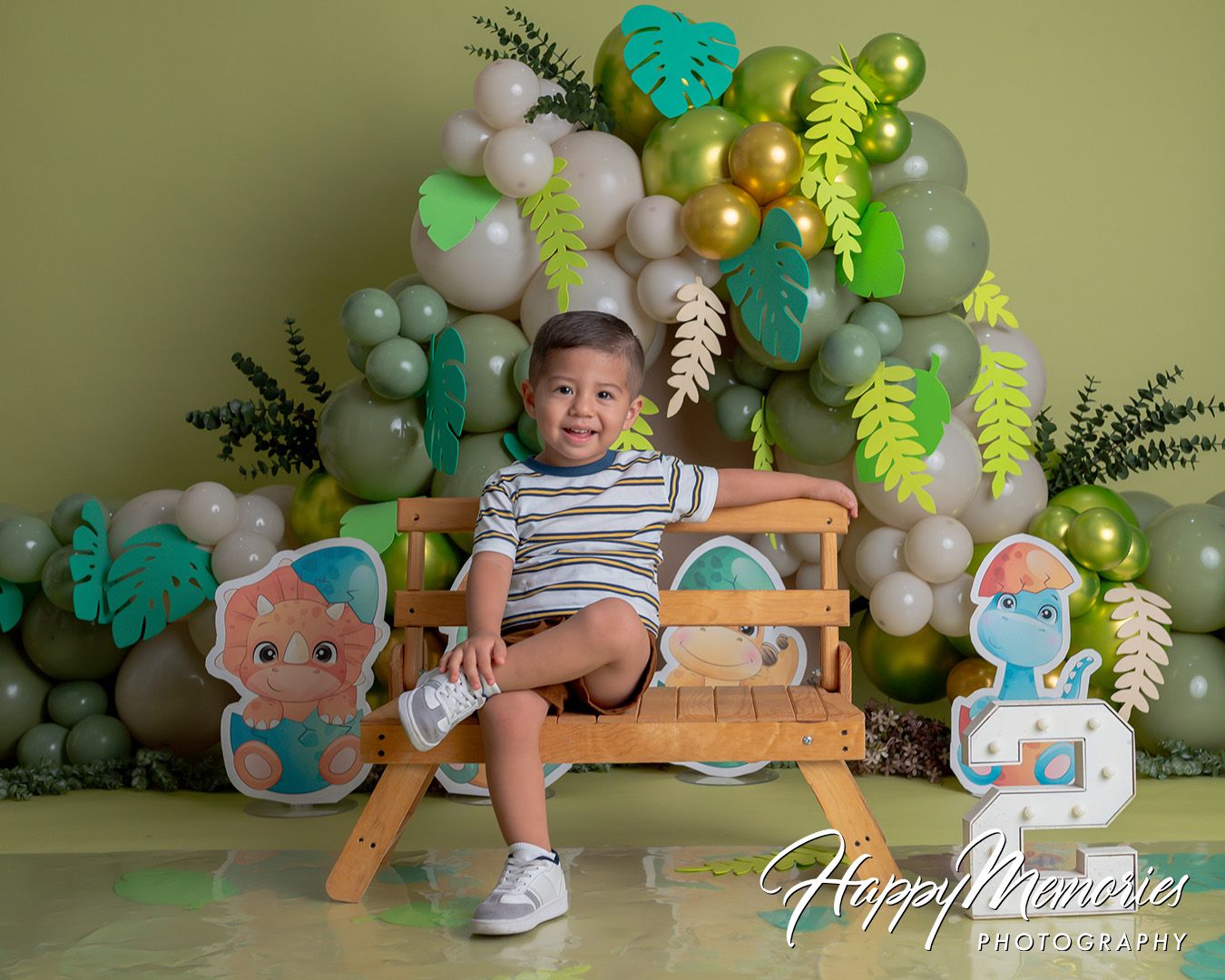 Toddler boy sitting on wooden bench with dinosaur cake smash decorations, green balloons, and tropical leaves backdrop.