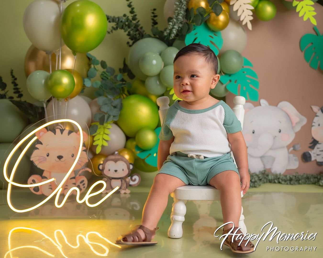 A first birthday party setup with green and gold balloon decorations, woodland theme decor, and cake smash photo session.