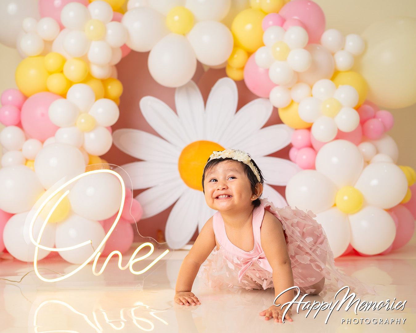 Baby photography setup with pink, yellow and white balloon arch featuring daisy decorations against neutral backdrop.