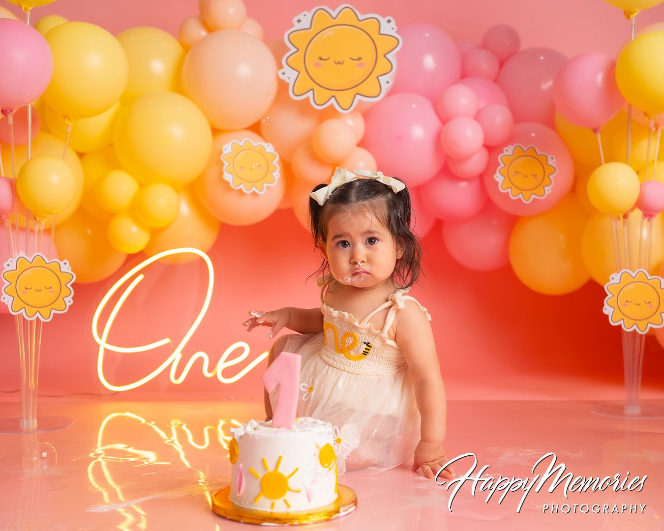 First birthday celebration with yellow and coral balloon arch, sunflower decorations, and a white cake on pink backdrop.