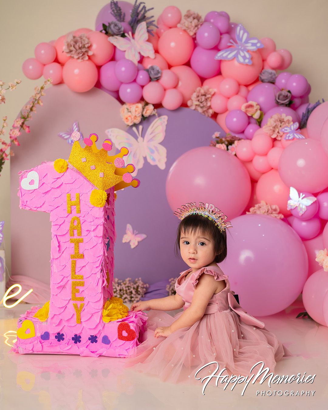 Pink and purple balloon arch with butterflies creates a dreamy backdrop for a first birthday celebration photoshoot.