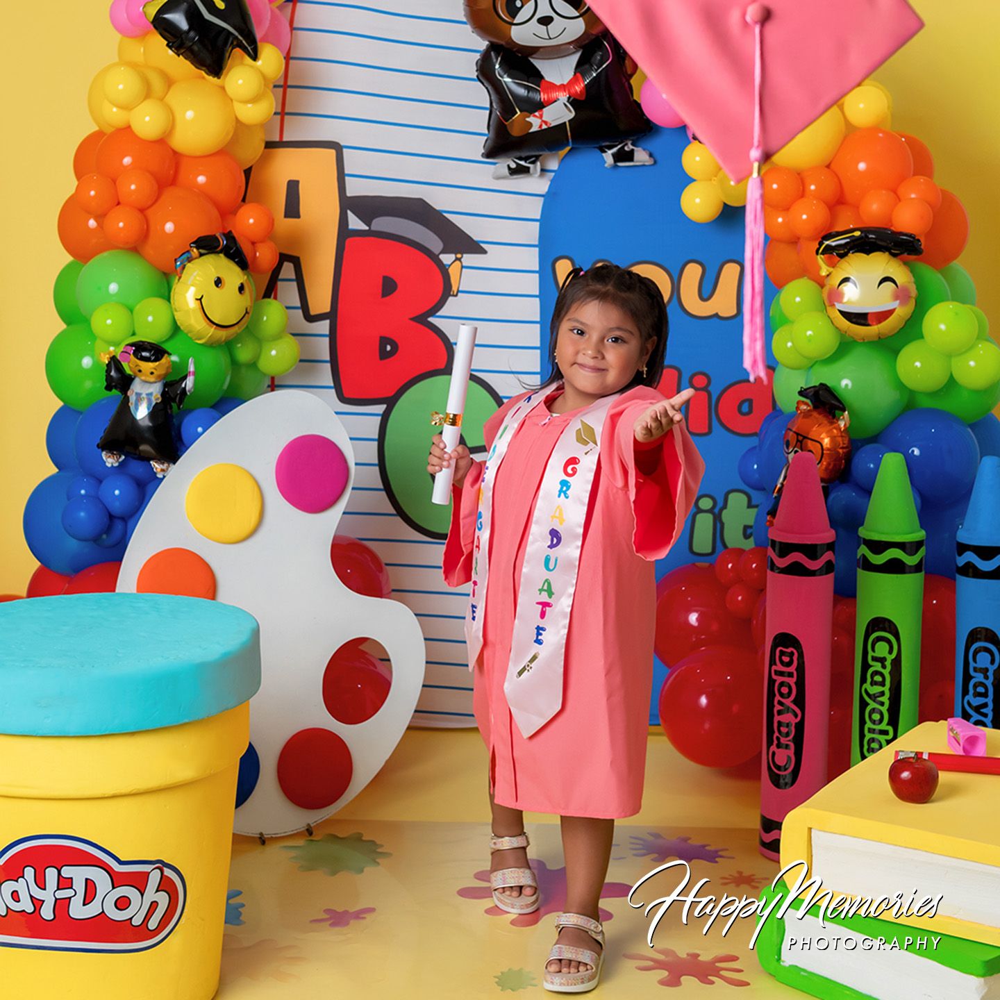 Young girl in pink graduation gown poses with diploma amid colorful balloon arch and Play-Doh decorations.