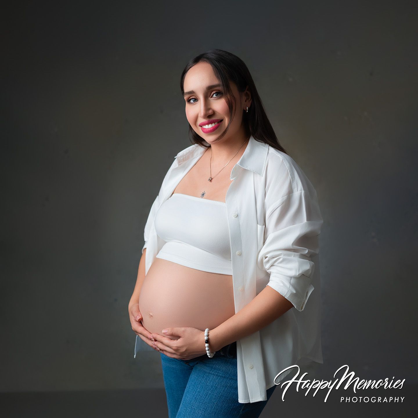 Smiling pregnant woman in white shirt and jeans cradling baby bump during professional maternity photo session.