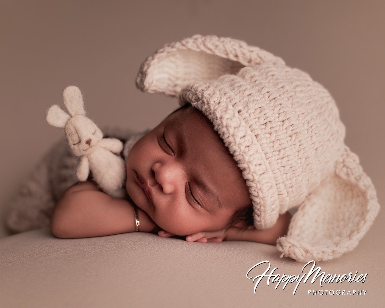 Sleeping newborn in cream knit hat cuddling small plush bunny toy during professional photo session.
