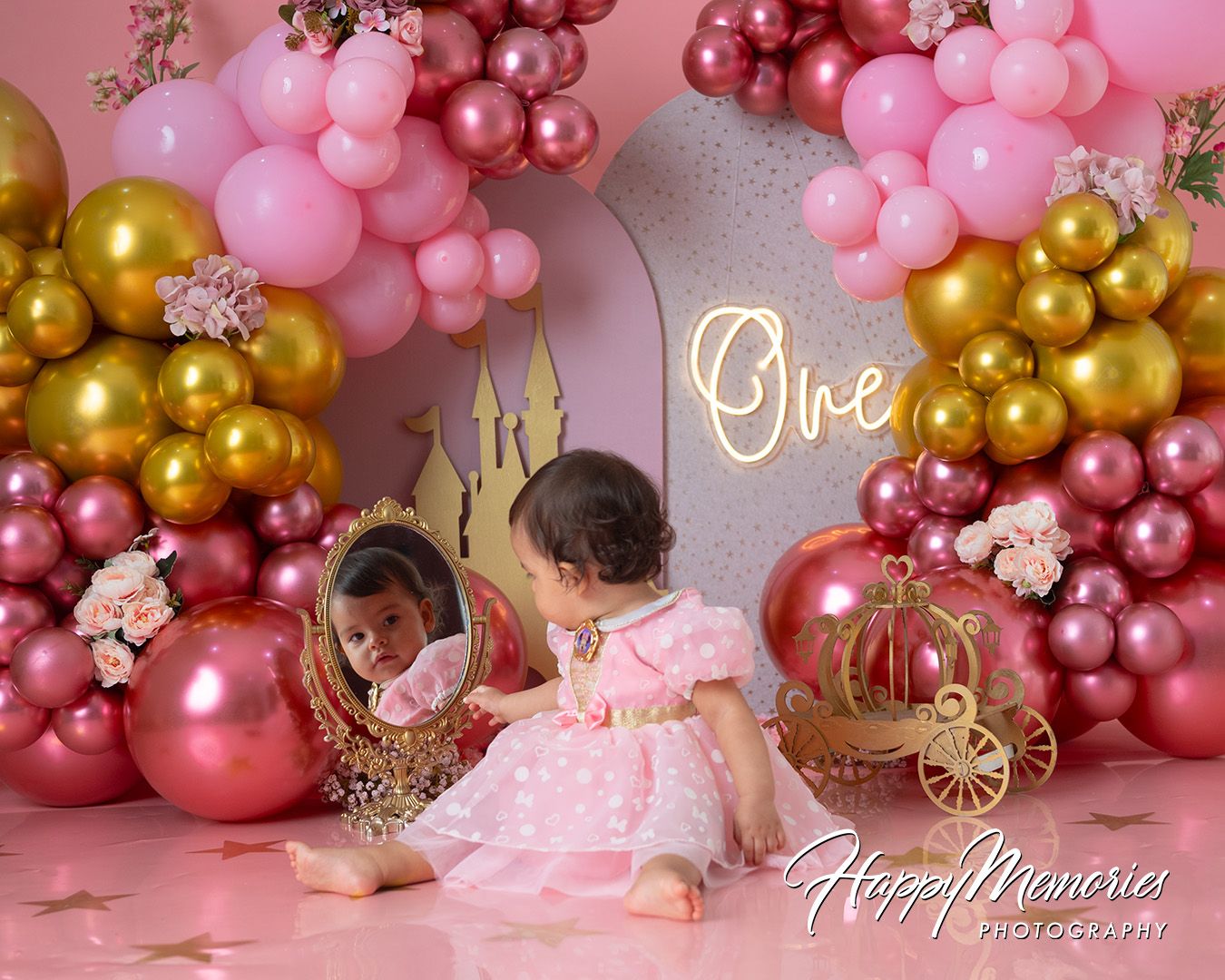 Baby girl in pink dress celebrates first birthday with gold and pink balloon arch and princess carriage decor.