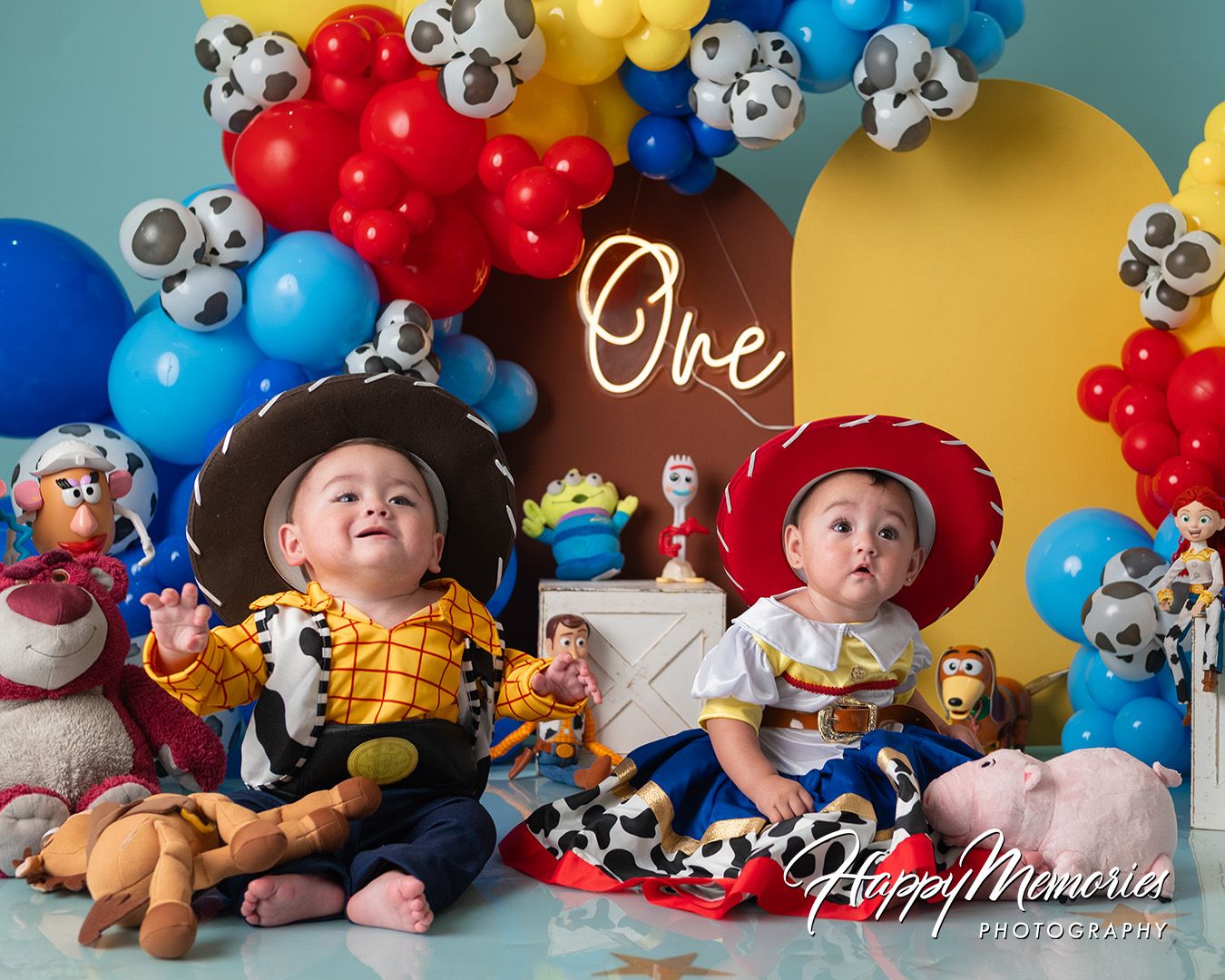 Twin babies dressed as Woody and Jessie from Toy Story celebrate their first birthday with colorful balloons and themed decorations.