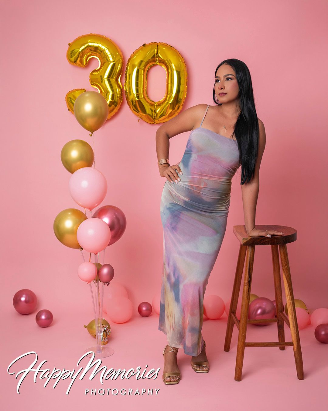 Birthday photoshoot with pink and gold balloons against pink backdrop featuring tie dye dress and wooden stool.