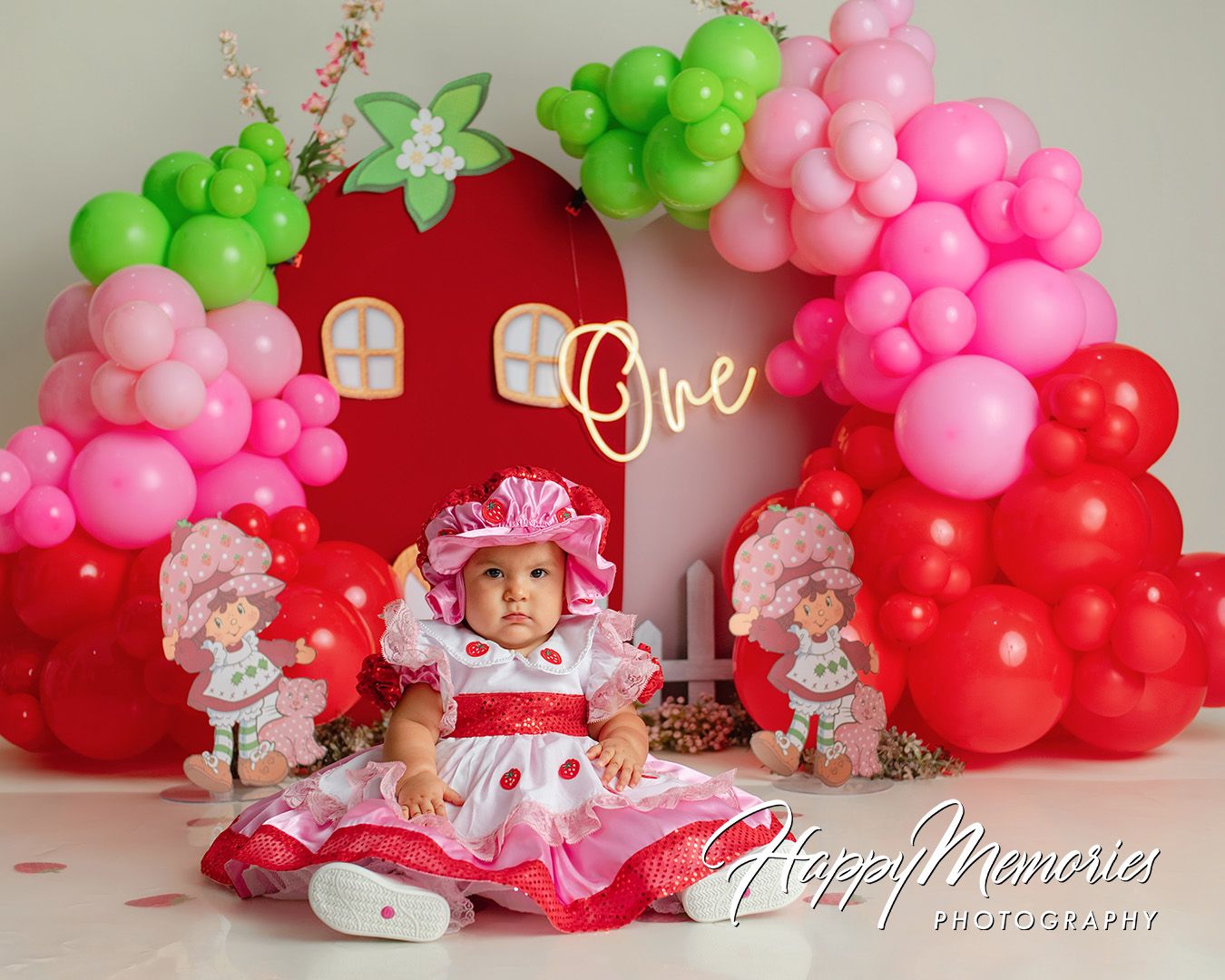 Baby girl in Strawberry Shortcake outfit sits before balloon arch and decorations at a first birthday cake smash.