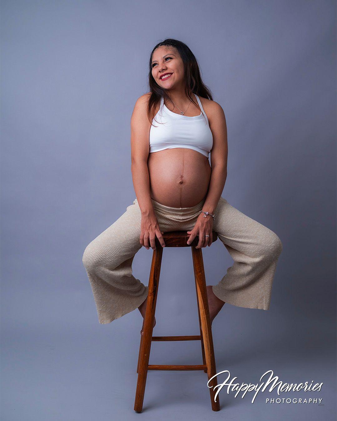 Studio maternity portrait with woman in white top and beige pants sitting on wooden stool.