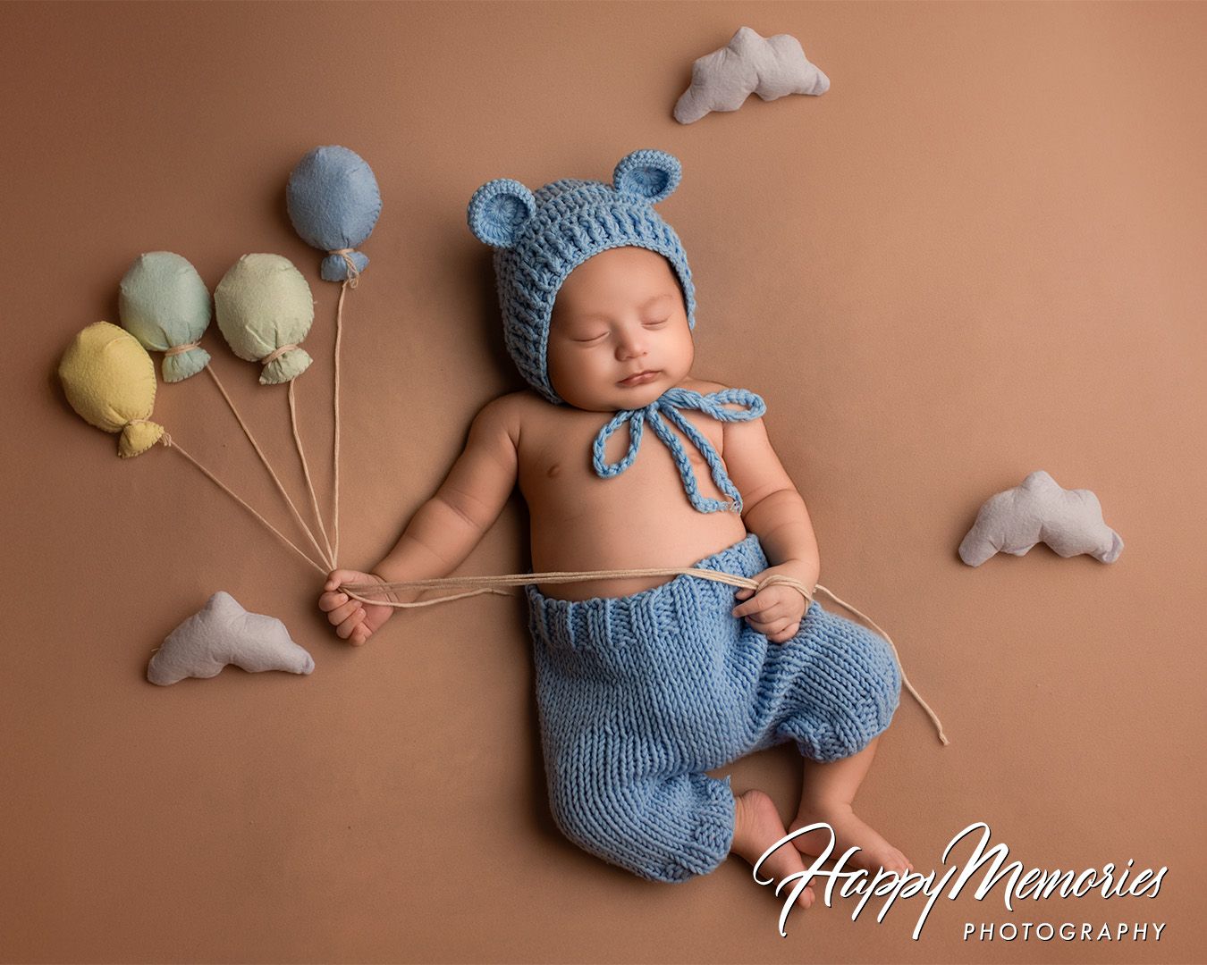 Newborn wearing blue knit outfit with adorable balloon props on brown background.