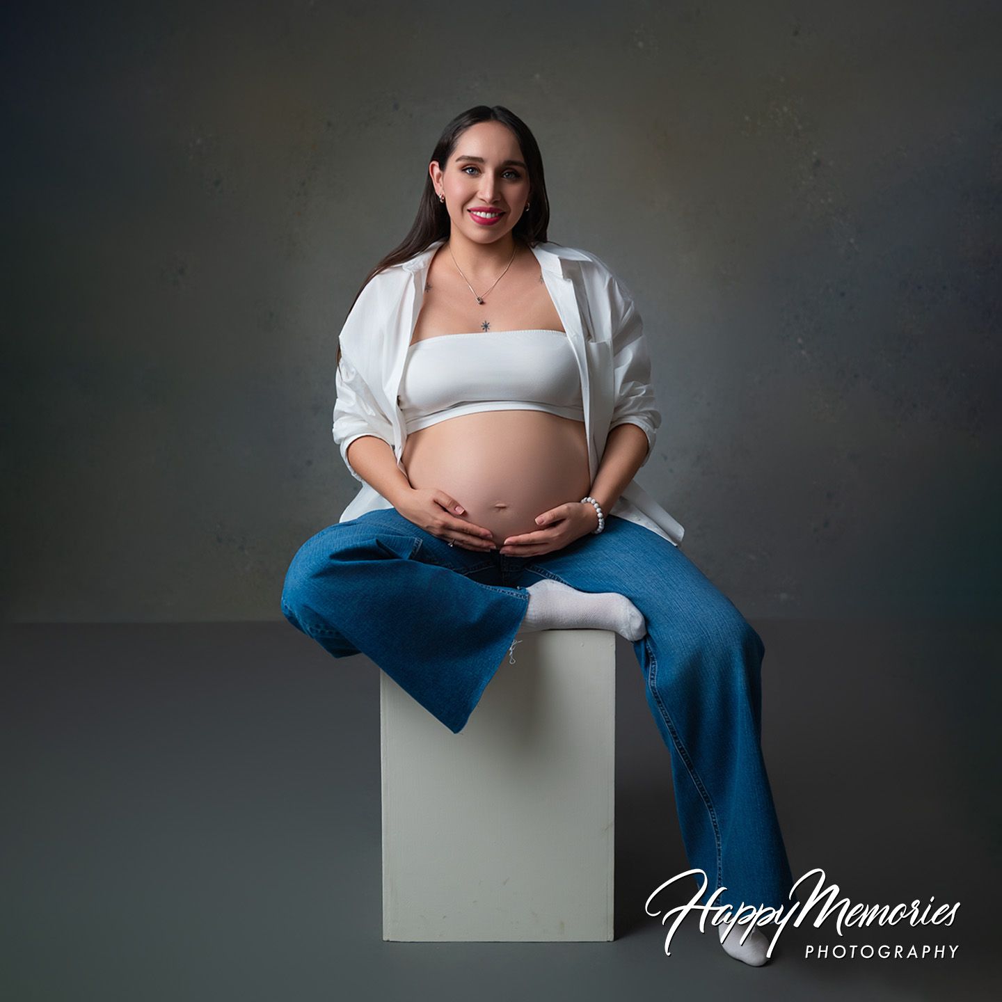 Pregnant woman in white crop top and blue pants cradling baby bump, seated on white cube for maternity photography session.