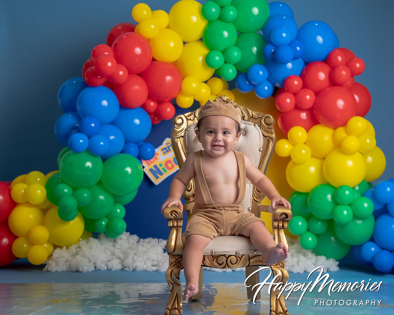 Baby in gold crown sits on ornate throne surrounded by colorful rainbow balloon arch for first birthday cake smash photo.