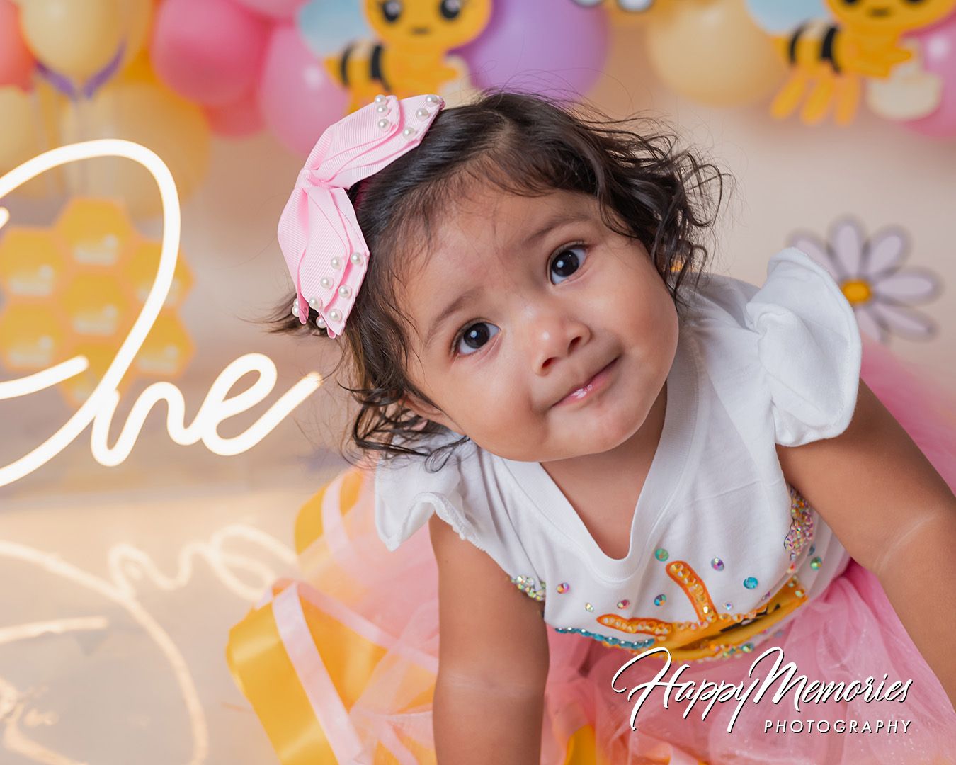 A child in a white dress with a pink bow poses against a colorful balloon backdrop with yellow cartoon decorations.
