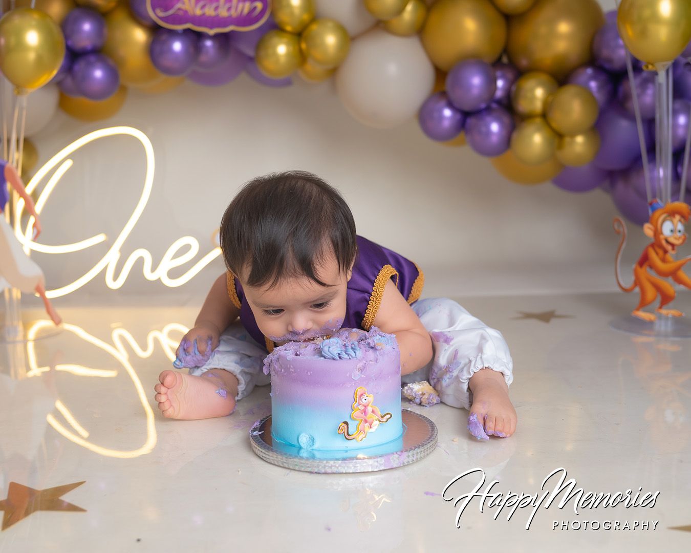 A birthday party setup with purple and gold balloon decorations and a light blue cake on a table.