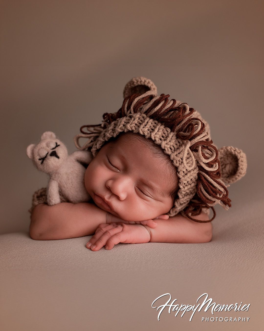 A sleeping newborn wearing a brown knit horse hat lies peacefully on a soft beige backdrop in a professional portrait setting.
